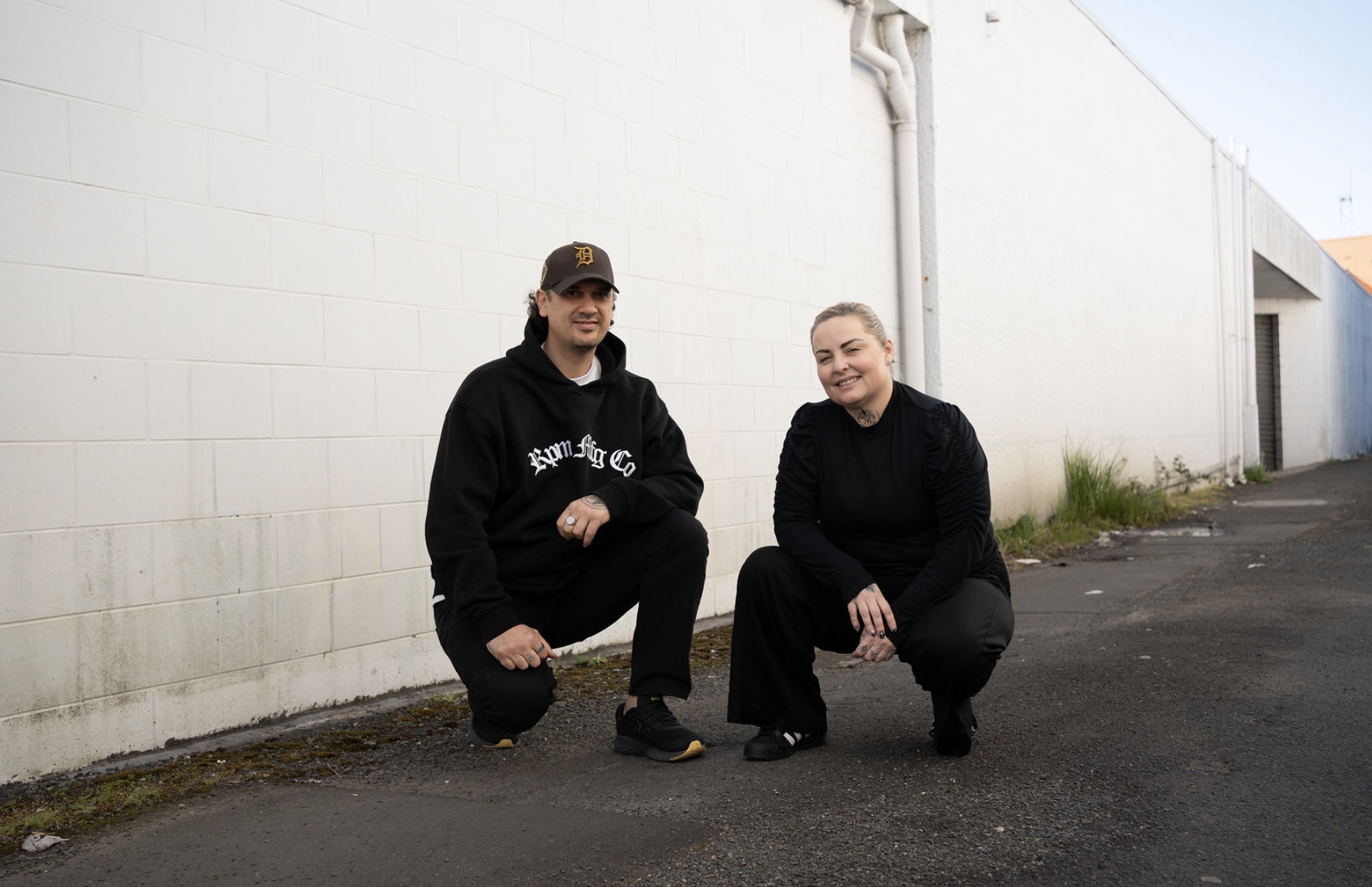 Cody Wharerau and Lisa Booth crouched against a white block wall in Kirikiriroa