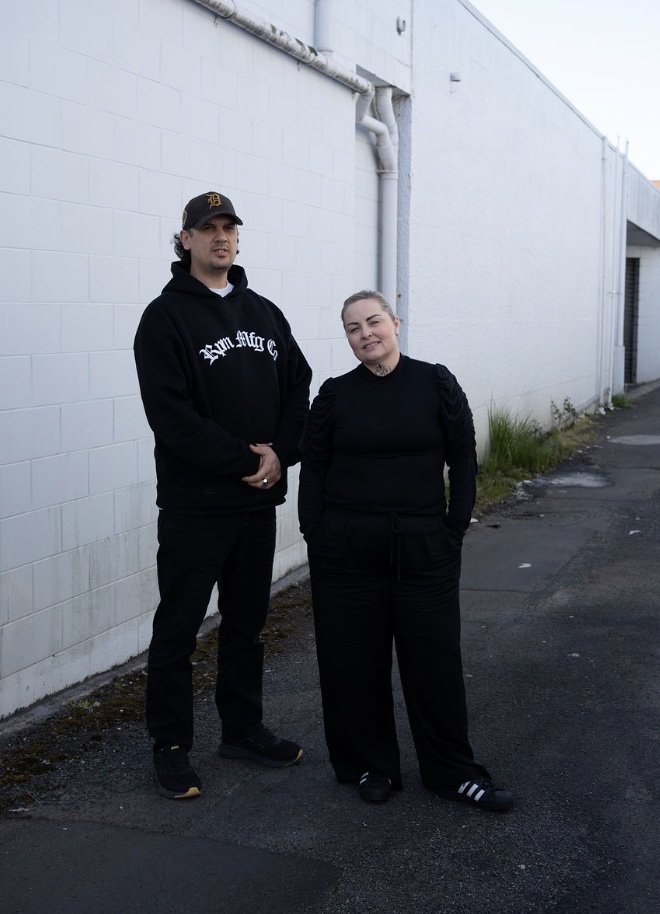 Cody Wharerau and Lisa Booth standing against a white block wall in Kirikiriroa